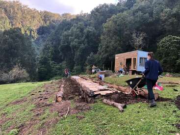 A group of people works outdoors, constructing a garden bed with logs and cardboard near a small wooden cabin, surrounded by lush green forest and hills.