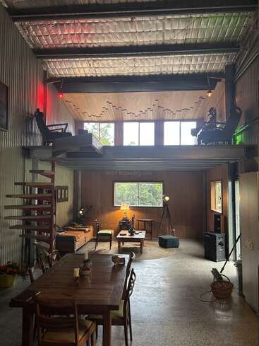 This industrial-style loft features a high ceiling, metal walls, exposed insulation, mezzanine workspace, cozy sitting area, wooden dining table, spiral stairs, and large windows.