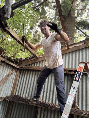 A man stands on a damaged wooden beam inside a shed, wearing safety glasses, gloves, and work clothes, holding a crowbar, smiling amid demolition.