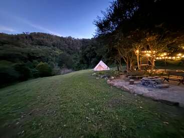 The image depicts a serene campsite at dusk, featuring a white tent, a wooden cabin with string lights, and a fire pit, set amidst a lush grassy area and trees.