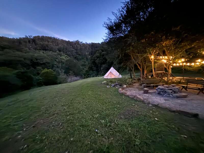 The image depicts a serene campsite at dusk, featuring a white tent, a wooden cabin with string lights, and a fire pit, set amidst a lush grassy area and trees.