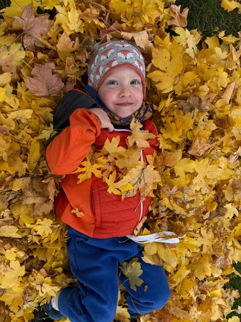 Un enfant souriant, vêtu d'une veste rouge et d'un chapeau à motif de renard, est allongé joyeusement sur le sol, entouré d'un lit vibrant de feuilles d'automne jaunes.