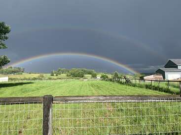 Um arco-íris duplo vibrante se estende sobre um campo verde exuberante, com uma cerca de madeira em primeiro plano e nuvens escuras de tempestade criando um contraste dramático.