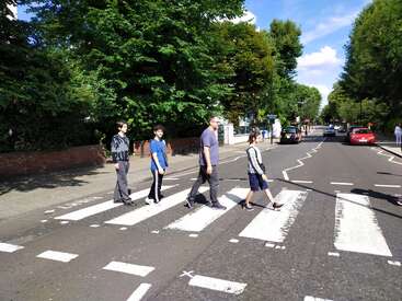 Cuatro personas cruzan un famoso paso de peatones en un día soleado, recreando la icónica escena de Abbey Road, rodeadas de árboles, edificios y coches.