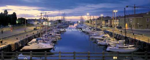 La imagen representa una serena escena de puerto deportivo al atardecer, con un canal bordeado de barcos y edificios, y una carretera que discurre por el lado izquierdo.