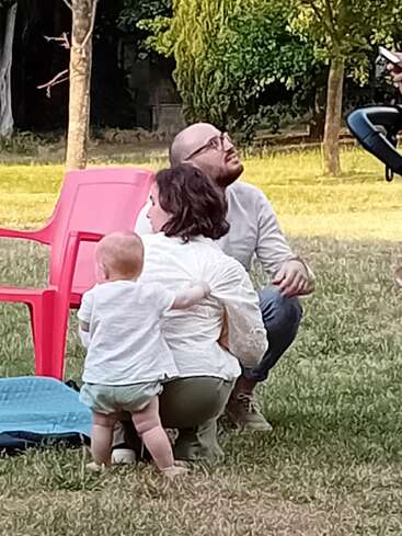 Un homme, une femme et un bébé profitent d'un moment en plein air sur de l'herbe. Une chaise rouge et un tapis bleu se trouvent à côté d'eux. Le bébé se tient debout, dans le dos de la femme.