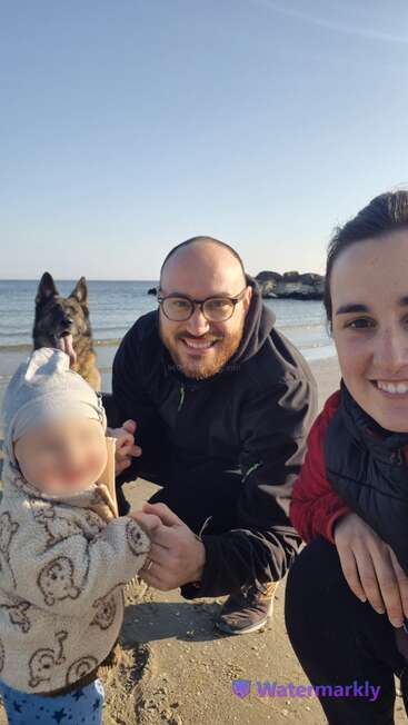 Una familia feliz, formada por un hombre, una mujer, un niño pequeño y un perro pastor alemán, posan juntos en una playa de arena con el mar de fondo.