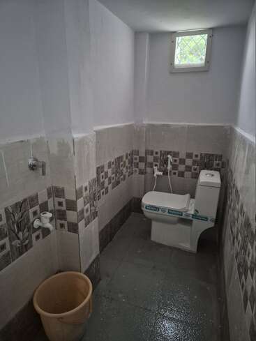 Cette image montre une petite salle de bains avec des toilettes blanches, des carreaux muraux à motifs, un jet d'eau, un seau, un robinet et une petite fenêtre grillagée.