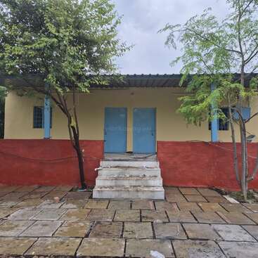 Cette image montre un bâtiment simple avec des murs jaunes, une partie inférieure rouge, des portes et des fenêtres bleues, deux arbres devant et des marches en pierre.