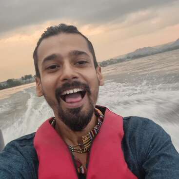 Un homme souriant portant un gilet de sauvetage rouge prend un selfie sur un bateau. L'eau clapote derrière lui, avec des montagnes et un ciel nuageux au coucher du soleil en vue.