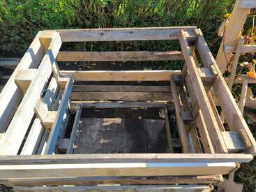 Cette image montre un simple bac à compost en bois fabriqué à partir de palettes en bois réutilisées, placé à l'extérieur dans un jardin avec des buissons verts à l'arrière-plan, sous la lumière du soleil.