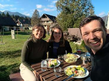Trois personnes souriantes prennent un repas en plein air à une table en bois dans un village pittoresque et ensoleillé avec des montagnes, de la verdure et de charmantes maisons en arrière-plan.
