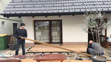Deux personnes construisent une terrasse en bois devant une maison. L'un soulève une planche de bois, tandis que l'autre, agenouillé, travaille avec des outils.