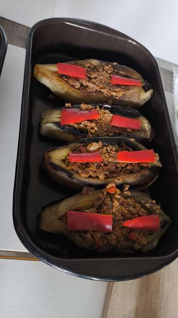 Trois aubergines cuites au four sont farcies d'un mélange de viande hachée et garnies de tranches de poivron rouge ; elles sont disposées avec soin dans un plat de cuisson noir, prêtes à être servies.