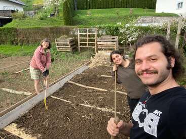 Trois personnes jardinent ensemble, souriantes, les mains occupées par des outils, dans un potager surélevé. À l'arrière-plan, on aperçoit de l'herbe verte, des arbres, des bacs à compost et des maisons. Un travail d'équipe en plein air.