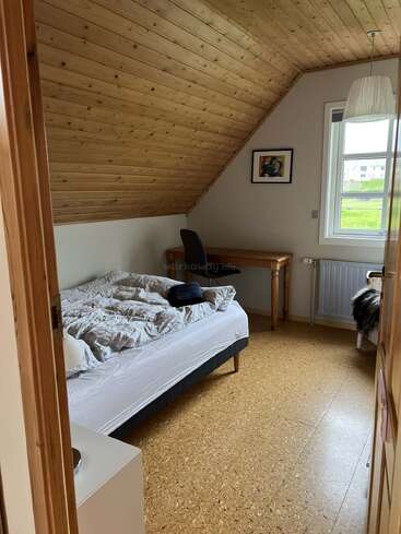 Cette chambre mansardée confortable dispose d'un plafond en bois incliné, d'un lit défait, d'un bureau avec une chaise, d'un tableau encadré, d'une fenêtre, d'une suspension et d'un sol en liège, créant une atmosphère chaleureuse.