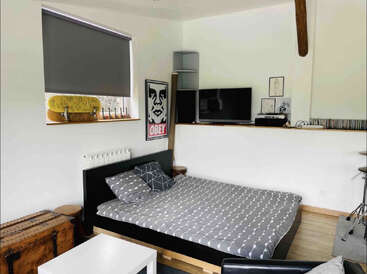 A modern bedroom with a grey bed, geometric bedding, TV, "Obey" poster, skateboard, drum set, wooden chest, and minimalist decor. Clean and stylish atmosphere.