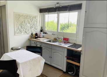 This image shows a bright, cozy kitchen with white cabinets, a table with a cloth, large window, dish rack, appliances, and wall art.
