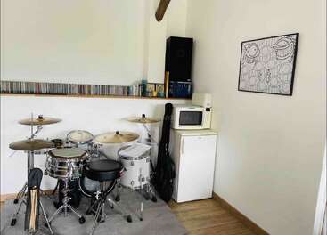 This cozy room features a drum set, a small fridge with a microwave on top, guitar cases, shelves with CDs, and abstract wall art.
