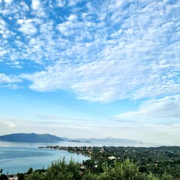 A beautiful coastal landscape features calm blue water, lush greenery, distant mountains, picturesque houses, and a stunning sky with scattered, fluffy white clouds on a bright day.