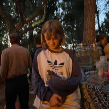 Una persona joven con una sudadera Adidas sostiene suavemente a un pequeño animal. La escena es al aire libre al atardecer, con gente, árboles y una mesa cerca.