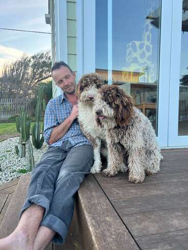 Un homme est assis, détendu, sous un porche en bois, à côté de deux grands chiens au poil frisé. Derrière eux se trouve une maison avec des portes vitrées et un jardin de cactus.
