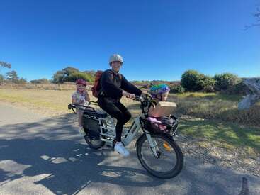 Un adulte souriant conduit un vélo électrique avec deux jeunes enfants, tous casqués, sur une route de campagne ensoleillée. Le ciel est clair et bleu.