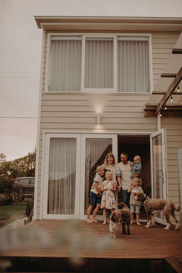 Une famille heureuse, composée de quatre enfants et de deux chiens, se tient sur une terrasse en bois à l'extérieur de leur maison moderne, souriante et profitant d'un moment de paix.