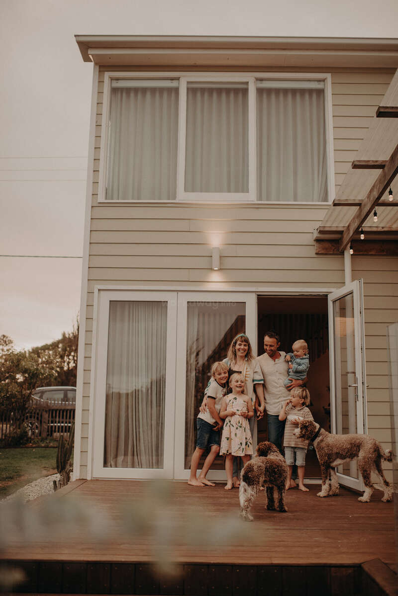 Une famille heureuse, composée de quatre enfants et de deux chiens, se tient sur une terrasse en bois à l'extérieur de leur maison moderne, souriante et profitant d'un moment de paix.