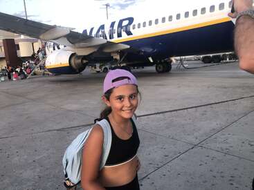 Une jeune fille souriante avec un sac à dos et une casquette violette se tient sur le tarmac d'un aéroport, avec en arrière-plan un avion de la compagnie Ryanair et des passagers qui embarquent. Le ciel est nuageux.