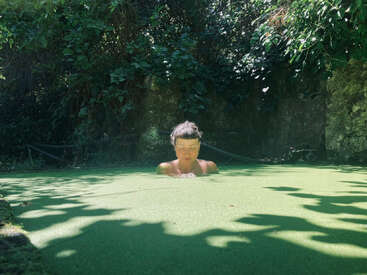 A imagem mostra uma mulher submersa em uma piscina de água, cercada por uma vegetação exuberante e um muro de pedra, com o rosto parcialmente obscurecido pela superfície da água.