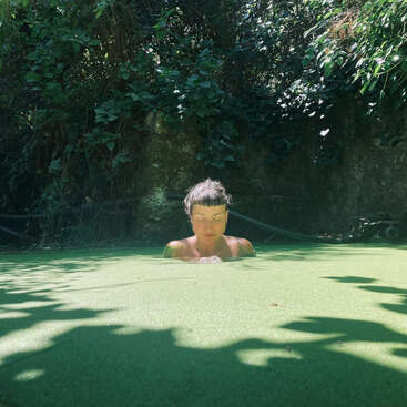 L'image représente une femme immergée dans un bassin d'eau, entourée d'une végétation luxuriante et d'un mur de pierre, dont le visage est partiellement masqué par la surface de l'eau.