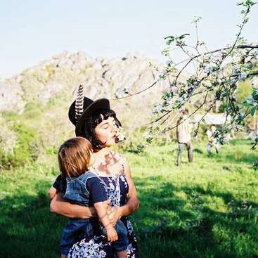The image depicts a woman holding a child in a grassy field, with a mountain in the background and a tree branch with white flowers in the foreground.