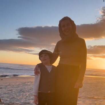 Eine Frau und ein Kind stehen zusammen an einem Sandstrand bei Sonnenuntergang. Das goldene Sonnenlicht beleuchtet sie und die Wellen des Meeres im Hintergrund.