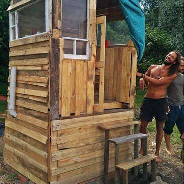 L'image représente une petite structure en bois avec une fenêtre et une porte, avec un homme debout à l'extérieur et une personne partiellement visible derrière lui, dans une zone boisée.