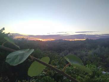 A imagem retrata um pôr do sol sereno em uma paisagem montanhosa, com vegetação exuberante em primeiro plano e um céu vibrante acima.