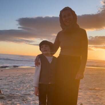 Una mujer y un niño están juntos en una playa de arena al atardecer, sonriendo con ternura. El cielo está lleno de nubes de colores y bañado por una suave luz dorada.