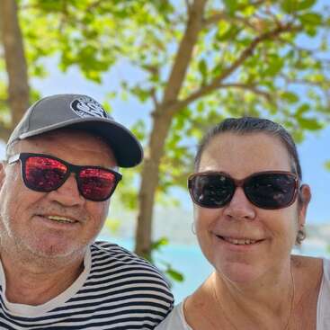 Un couple âgé, tout sourire, portant des lunettes de soleil, pose en plein air devant des arbres aux feuilles vertes. L'homme porte une chemise rayée et une casquette ; tous deux ont l'air heureux et détendus.