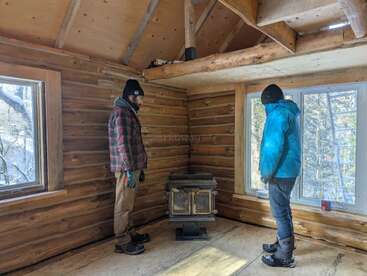 Two people in winter clothing stand inside a rustic wooden cabin, observing a wood stove. Sunlight streams through large windows, illuminating the cozy log interior.