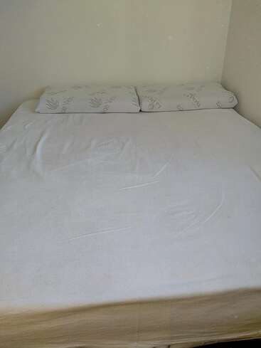 Das Bild zeigt ein ordentlich gemachtes Bett mit einem weißen Spannbettlaken und zwei Kissen, das an einer schlichten weißen Wand in einer Zimmerecke steht.
