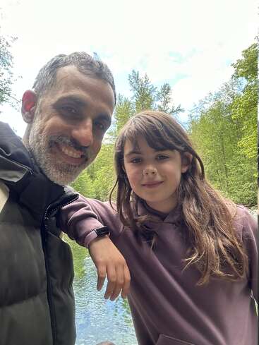 La imagen muestra a un hombre y una niña posando para un selfie frente a una serena masa de agua, rodeados de frondosos árboles verdes y un cielo luminoso.