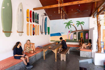 Une pièce à l'ambiance décontractée sur le thème de la plage, avec des planches de surf et de skate accrochées au mur, une fresque représentant des palmiers, et des personnes discutant sur des bancs encastrés autour d'une table en bois.