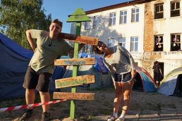 An einem sonnigen Tag stehen zwei Menschen lächelnd neben einem bunten Holzschild mit Begrüßungen in mehreren Sprachen, vor Zelten und einem alten Gebäude.