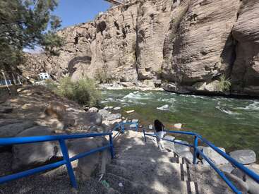 Eine Person steigt eine Steintreppe mit blauem Geländer hinunter zu einem klaren, schnell fließenden Fluss. Dahinter erheben sich schroffe Felsen, Bäume und Sonnenlicht schaffen eine friedliche, natürliche Schönheit.