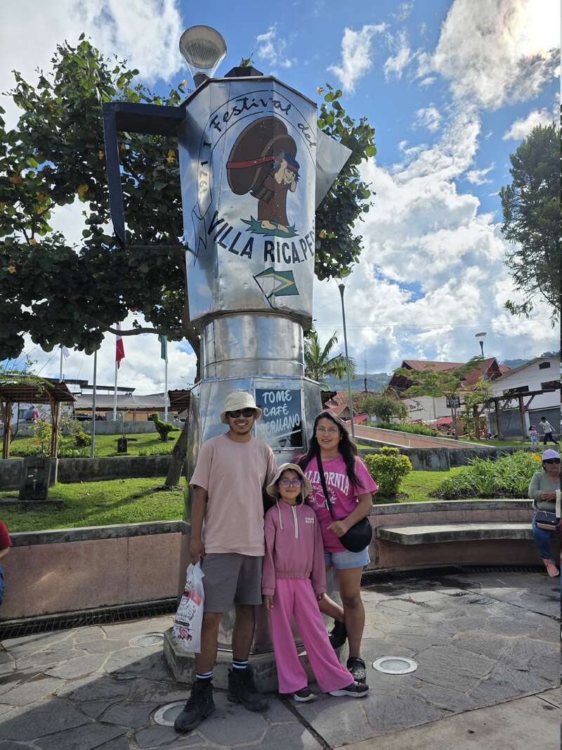 Eine dreiköpfige Familie posiert lächelnd vor einem großen metallischen Kaffeekannen-Denkmal mit der Aufschrift "Villa Rica". Im Hintergrund sind ein sonniger Platz und Grünflächen zu sehen.