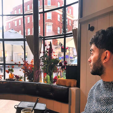 A man with a beard sits indoors, gazing out a large paned window. Cozy cafe atmosphere, flowers in vases, city street and brick building outside.