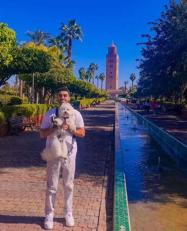 A man in white clothes holds a fluffy white dog near a fountain, with palm trees, benches, and a tall historic tower under a bright blue sky.