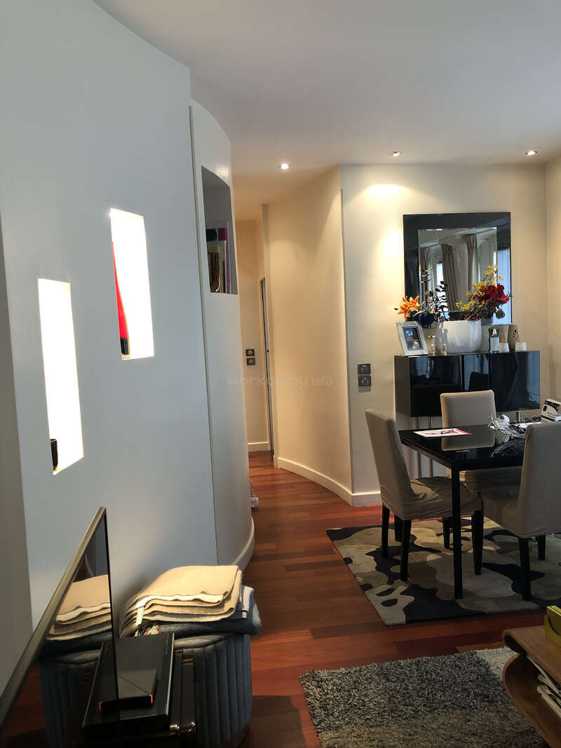 L'image montre une pièce moderne et bien éclairée avec un coin repas, une table et des chaises, un miroir et un couloir menant à une autre pièce, avec une pile de couvertures pliées sur le sol.