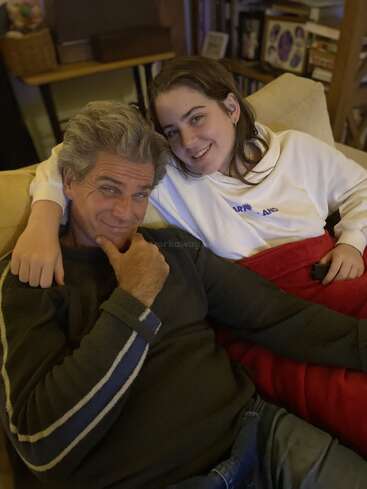 A man and a young woman sit closely together on a couch, smiling warmly. The atmosphere feels cozy and relaxed, suggesting a close, happy relationship.
