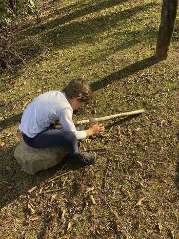 A young boy sits on a rock outdoors, intently playing with sticks on the ground, surrounded by grass, sunlight, and scattered leaves, enjoying nature.